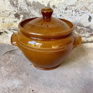 May include: A brown ceramic casserole dish with a lid. The dish has two handles and a decorative dotted pattern around the rim. The lid has a small knob and a vent hole. The dish is sitting on a stone surface.