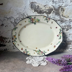 May include: An oval ceramic serving platter with a cream-coloured base and a dark rim. The border features a repeating pattern of small, colourful floral clusters in red, yellow, and green, with dark brown branches connecting the blossoms.