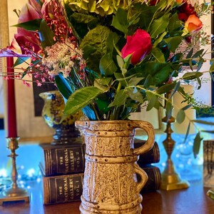 May include: A beige ceramic jug with a handle, holding a colourful floral arrangement. The bouquet features red, orange, and pink roses, green hydrangeas, and other flowers. The jug is placed on a wooden surface.