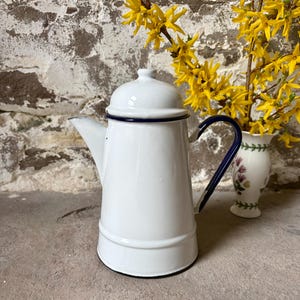 May include: White enamel coffee pot with a dark blue handle and trim. The vintage-style pot has a lid and spout. A small vase with yellow flowers is in the background.