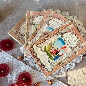 May include: Several antique books titled "History of Everyday Things in England" with illustrations. The books have a pink and cream color scheme, with decorative borders and text. They are arranged on a white lace doily, with red floral decorations.