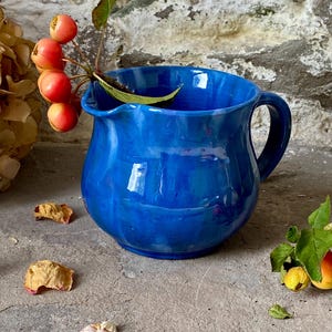 May include: A bright blue ceramic jug with a handle and a pouring spout. The jug has a glossy finish. A sprig of red berries and green leaves provides a natural accent. The jug is likely used for serving drinks.