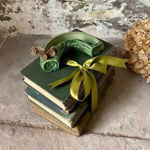 May include: A collection of vintage green books is decorated with a green ceramic ornament and a green ribbon. The ceramic piece features a curved design with a floral pattern. Dried flowers add a touch of natural elegance to the composition.