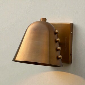 May include: A bronze-colored wall sconce with a bell-shaped shade and a rectangular backplate. The shade has a rounded top and a series of small, round rivets. The backplate has the text "DualLighting786" printed on it.