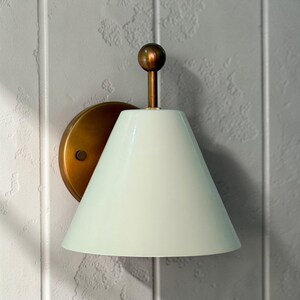 May include: A wall-mounted light fitting featuring a brass-coloured circular base and a conical, off-white shade. The fitting has a small, round, brass-coloured accent at the top.