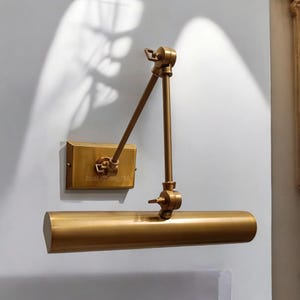 Może przedstawiać: Mosiężna lampa ścienna z regulowanym ramieniem i cylindrycznym kloszem. Na prostokątnej płycie ściennej widnieje napis "DualLighting786". Idealna do oświetlania obrazów lub dzieł sztuki.