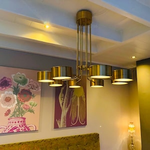 May include: A modern gold-toned chandelier with multiple cylindrical shades illuminates a room. The fixture hangs from the ceiling, complemented by artwork featuring floral arrangements. The room's decor includes a painting with pink and green flowers.