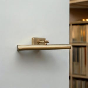 May include: A brass-coloured picture light fixed to a white wall. The light features a rectangular base and a long, cylindrical arm. The fixture includes a small, adjustable knob and a rectangular base.