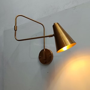 Peut inclure: Une lampe murale en laiton avec un abat-jour conique. La lampe a un bras rectangulaire et une base circulaire. La lumière est allumée, dégageant une lueur chaude. Le texte "DuaLighting786" est visible sur le mur.
