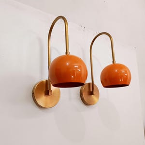 May include: Two wall lamps with orange globe shades and curved brass arms. The lamps have circular brass bases and are mounted on a white wall. The shades have a glossy finish. The design is modern, suitable for interior decor.