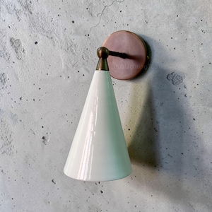 May include: A white cone-shaped wall sconce with a bronze-colored mounting plate and a brass-colored arm. The sconce is mounted on a textured, light gray wall. The sconce has a glossy finish and a modern design.
