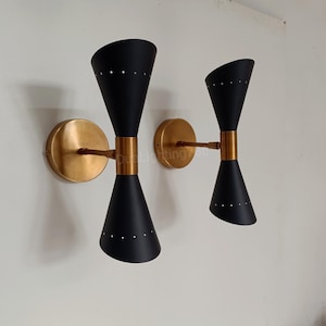 Peut inclure: Deux appliques murales modernes du milieu du siècle. Chaque applique a un abat-jour noir en forme de sablier avec de petits points blancs espacés. Les appliques ont une base circulaire dorée et une bande centrale dorée.