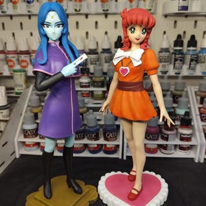 Może przedstawiać: Dwie figurki w stylu anime na oddzielnych podstawkach. Lewa postać ma niebieskie włosy, jasnoniebieską skórę i fioletową sukienkę z czarnymi butami. Prawa postać ma rude włosy, pomarańczową sukienkę z białą kokardą i czerwone buty na podstawce w kształcie serca.