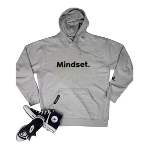 Op de afbeelding: Een grijs hoodie met het woord "Mindset." in zwart gedrukt op de voorkant. De hoodie heeft een zak op de voorkant en een kleine "M." gedrukt op de linkermouw. Een paar zwart-witte Converse sneakers liggen op de vloer voor de hoodie.