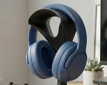 May include: A pair of blue over-ear headphones rests on a black headphone stand. The stand has a curved design and a wide base. A black game controller sits on the wooden desk in the foreground.