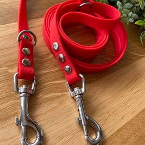 May include: A bright red double dog lead with two silver clips. The lead is made of a durable, rubber-like material.