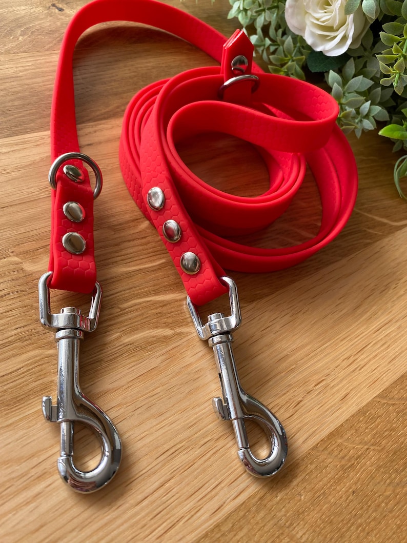 May include: A bright red double dog lead with two silver clips. The lead is made of a durable, rubber-like material.