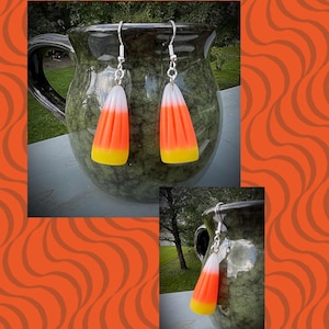 May include: A pair of Halloween-themed earrings shaped like candy corn. Each earring features a white, orange, and yellow design, suspended from silver-colored hooks. The earrings are displayed against a green ceramic mug and an orange background.