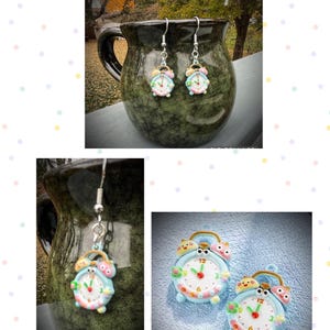 May include: A pair of whimsical clock earrings. Each earring features a light blue clock face with a white center, red hands, and a small, smiling owl on top. The earrings are suspended from silver-colored hooks.