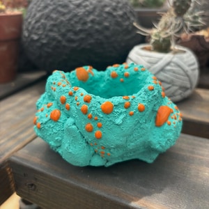 May include: A turquoise-colored, textured ceramic planter with orange polka dots. The small, round pot has an irregular shape and is designed for small plants. Other planters are visible in the background.
