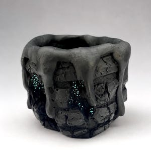May include: A small, dark gray, textured pot with a rough, brick-like exterior surface. Thick, smooth, matte black glaze drips down from the rim over the sides. Small, sparkling, teal-colored crystalline accents are visible within the crevices.