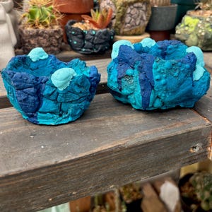 May include: Two small, textured planters with a rugged, uneven surface. The planters feature a layered color palette of deep navy blue, bright turquoise, and accents of pale cyan, creating a rocky, organic appearance with a hollow center.