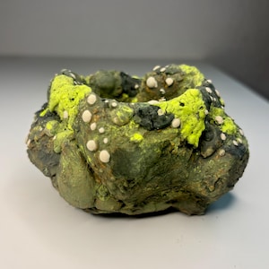 May include: A small, irregular, bowl-shaped object with a textured, earthy green and dark gray surface. The rim is accented with vibrant neon yellow patches and small, raised white circular dots scattered across the uneven, organic exterior.
