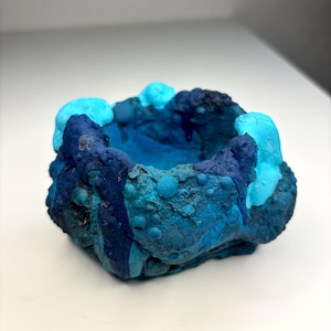 May include: A small, bowl-shaped object with a highly textured, uneven surface. The exterior features a gradient of blue tones, ranging from deep navy to bright cyan, with raised, bubble-like protrusions and drips of darker blue paint.
