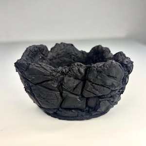 May include: A small, matte black bowl with a highly textured, craggy exterior surface that resembles broken rock or volcanic stone. The rim is uneven and jagged, featuring deep crevices and sharp, angular protrusions throughout the entire vessel.