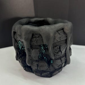 May include: A black, textured, cylindrical container with a thick, dripping glaze effect around the top rim. The exterior features a brick-like pattern with small, recessed gaps that reveal a glowing, speckled teal-colored interior surface.