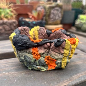 May include: A small, round, hand-sculpted planter with a rough, uneven texture. The exterior features a multi-colored design with patches of matte black, bright yellow, and vibrant orange paint applied over a muted, earthy brown base.