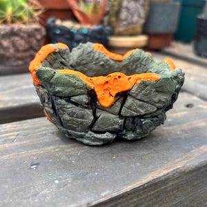 May include: A small, handcrafted succulent planter with a unique, textured design. The planter is primarily gray with an orange rim, giving it a volcanic rock appearance. The interior is unglazed, and the planter sits on a wooden surface. Other planters are visible in the background.