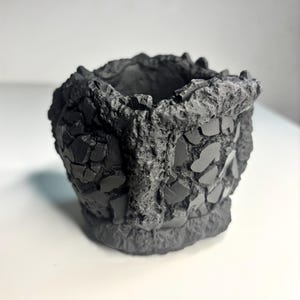May include: A small, dark gray, cylindrical vessel with a rough, craggy exterior texture. The surface features a mosaic-like pattern of flat, smooth, dark gray shards embedded within a coarse, uneven, rock-like material around the rim and base.