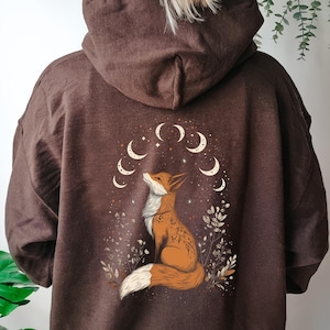 May include: A brown hoodie with a graphic of a fox looking up at a crescent moon and stars. The fox is surrounded by small flowers and leaves.