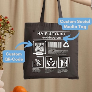 Op de afbeelding: Donkergrijze draagtas met de tekst "HAIR STYLIST @addcostum" en een QR-code. De tas heeft iconen van scharen, harten en een persoon die in een spiegel kijkt. De tas heeft ook een lijst met ingrediënten.