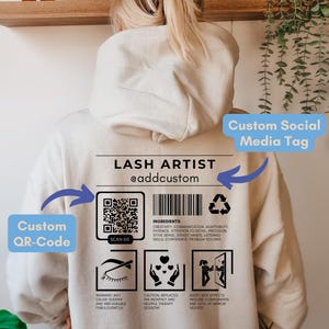 May include: A beige hoodie with a black and white graphic design that says "Lash Artist @addcustom". The graphic includes a QR code, a list of ingredients, and three icons. The text "Custom QR-Code" and "Custom Social Media Tag" are written in blue boxes on the image.
