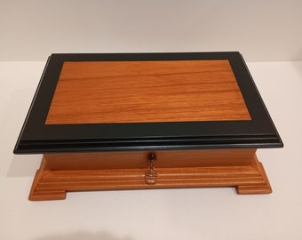 May include: A wooden box with a black trim and a small drawer. The box is made of light wood with a reddish hue. The drawer has a small metal knob.