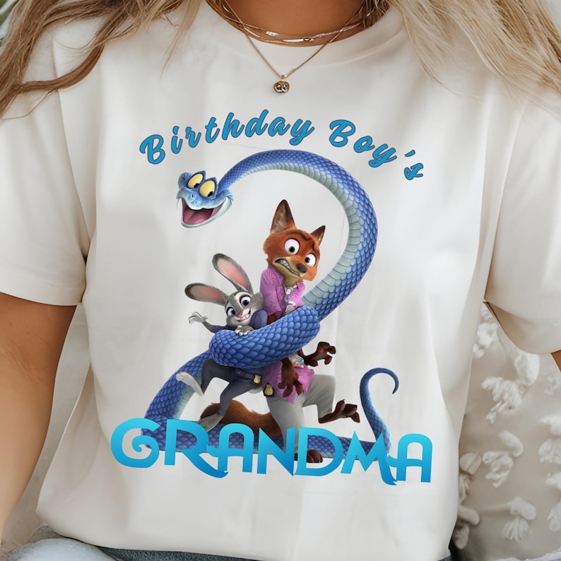 May include: A white t-shirt featuring a graphic of a blue snake coiled around a fox and a rabbit. The text "Birthday Boy's" is printed in blue above the characters, and the word "GRANDMA" is printed in blue below the characters.