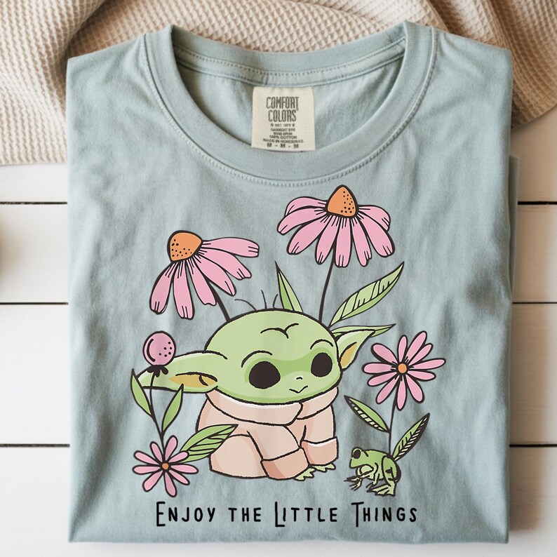 May include: A light blue t-shirt featuring a graphic of a green creature with large ears, surrounded by pink flowers and a small green frog. The text "ENJOY THE LITTLE THINGS" is printed in black below the illustration. The shirt has a Comfort Colors label.