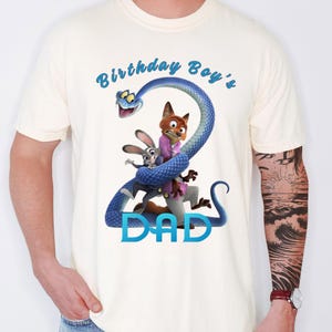 May include: A cream-colored short-sleeve t-shirt featuring a graphic of a blue snake coiled into the shape of the number two, with cartoon characters Judy Hopps and Nick Wilde. The text reads Birthday Boy's at the top and DAD at the bottom.