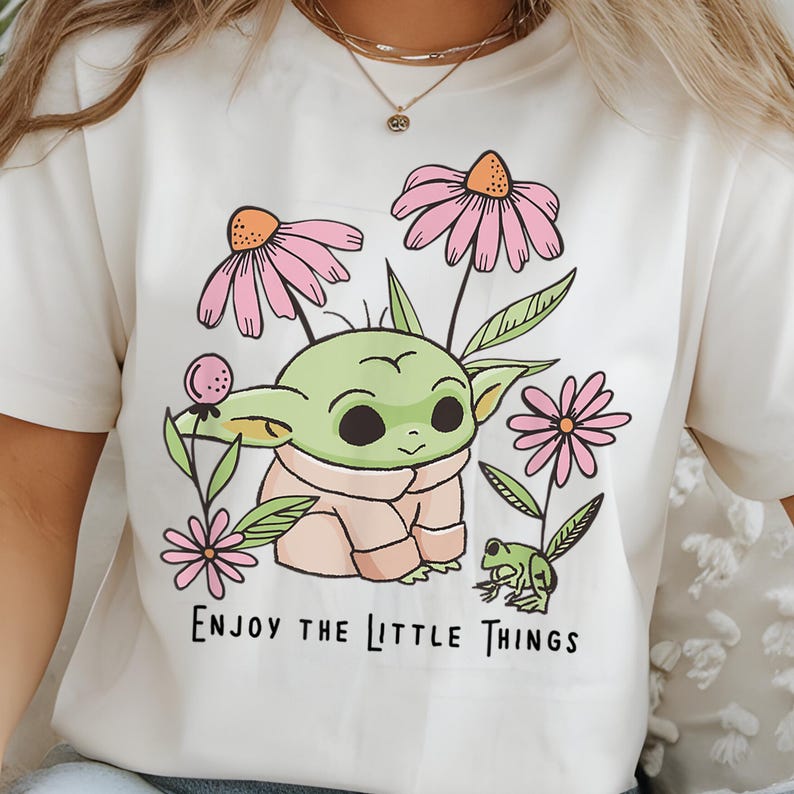 May include: A cream-colored t-shirt featuring a graphic illustration of a green creature with large ears, surrounded by pink flowers and a small green frog. The text "ENJOY THE LITTLE THINGS" is printed in black below the central illustration.