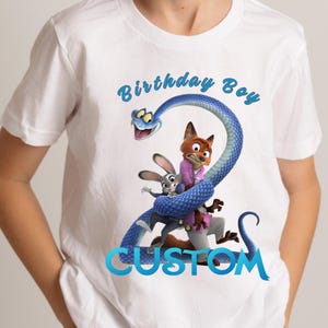 May include: A white short-sleeved t-shirt featuring a graphic of a blue snake coiled into the shape of the number two, with a cartoon rabbit and fox character inside the coil. The text Birthday Boy is printed in blue above, and CUSTOM below.