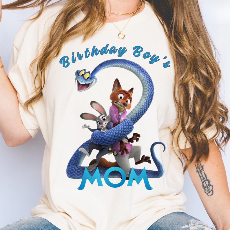 May include: A cream-colored t-shirt featuring a graphic of a blue snake forming the number two, with a cartoon fox and rabbit character positioned in the center. The text Birthday Boy's is printed above, and the word MOM is printed below in blue.