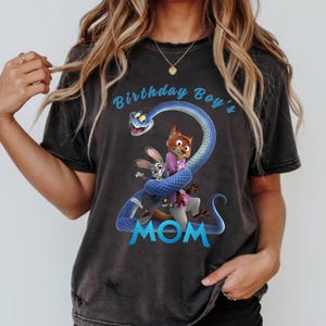 May include: A charcoal grey short-sleeve t-shirt featuring a graphic of a blue snake coiled into the shape of the number two, with cartoon characters Judy Hopps and Nick Wilde. The text reads Birthday Boy's MOM in light blue lettering.