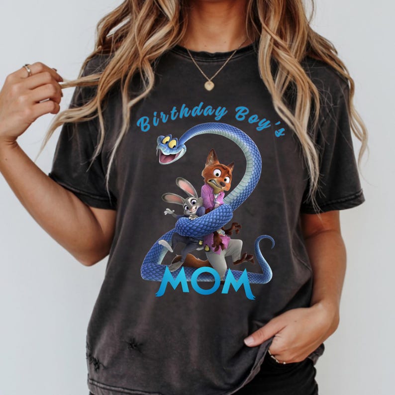 May include: A charcoal grey short-sleeve t-shirt featuring a graphic of a blue snake coiled into the shape of the number two, with cartoon characters Judy Hopps and Nick Wilde. The text reads Birthday Boy's MOM in light blue lettering.