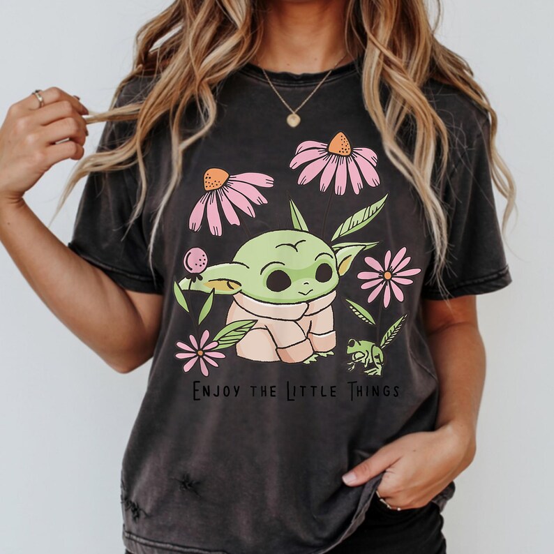 May include: A charcoal gray short-sleeve t-shirt featuring a graphic of a green creature with large ears, surrounded by pink flowers and green leaves. The text "ENJOY THE LITTLE THINGS" is printed in black below the central illustration.