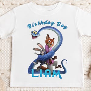 May include: A white short-sleeve t-shirt featuring a graphic of a blue snake coiled into the shape of the number two, with a cartoon fox and rabbit character inside the coils. The text reads Birthday Boy at the top and LIAM at the bottom.