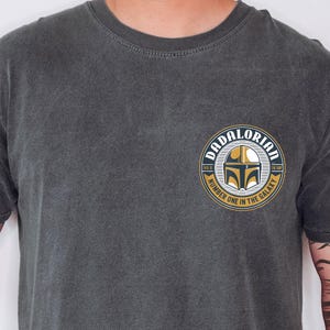 May include: A charcoal gray crew neck t-shirt featuring a circular graphic on the left chest. The design displays a gold and black helmet icon with the text DADALORIAN, THIS IS THE WAY, and NUMBER ONE IN THE GALAXY in a circular border.