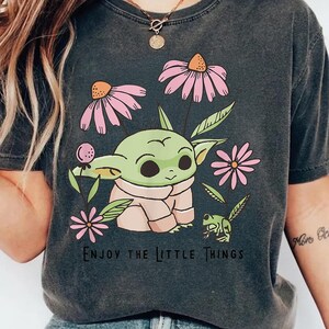 May include: A charcoal gray t-shirt featuring a graphic of a green creature with large ears, surrounded by pink flowers and a small green frog. The text "ENJOY THE LITTLE THINGS" is printed in black below the illustration on the front of the shirt.