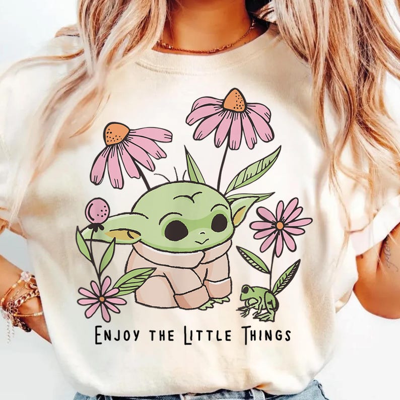 May include: A cream-colored t-shirt featuring a graphic illustration of a small green creature with large ears, surrounded by pink flowers and a small green frog. The text "ENJOY THE LITTLE THINGS" is printed in black below the illustration.